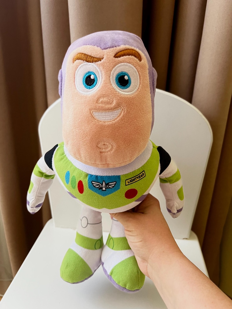 May include: A plush Buzz Lightyear doll, featuring a light purple head and a green and white spacesuit. The doll has large blue eyes, a smiling expression, and the word "LIGHTYEAR" on the chest. The doll is held in front of a white chair.