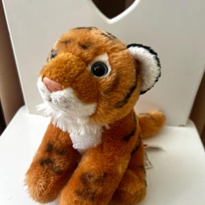 Op de afbeelding: Een pluche tijgerwelp met oranje en zwarte strepen, witte vacht rond de nek en een roze neus. Het knuffeldier heeft grote, donkere ogen en zit rechtop. Er is een label aan bevestigd.