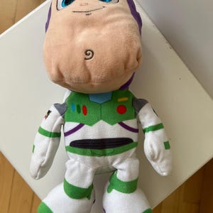 Pode incluir: Um boneco de pel&uacute;cia do Buzz Lightyear da franquia Toy Story. O boneco apresenta uma cabe&ccedil;a roxa clara, um rosto cor de p&ecirc;ssego e um traje espacial verde e branco. O boneco tem olhos azuis grandes e uma express&atilde;o amig&aacute;vel.