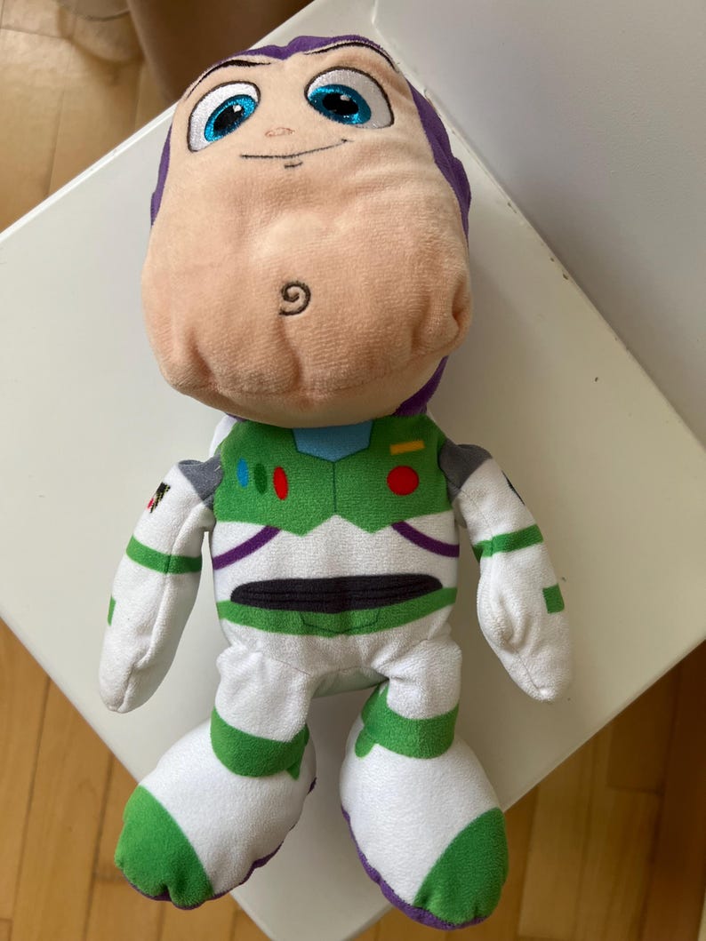 Pode incluir: Um boneco de pel&uacute;cia do Buzz Lightyear da franquia Toy Story. O boneco apresenta uma cabe&ccedil;a roxa clara, um rosto cor de p&ecirc;ssego e um traje espacial verde e branco. O boneco tem olhos azuis grandes e uma express&atilde;o amig&aacute;vel.