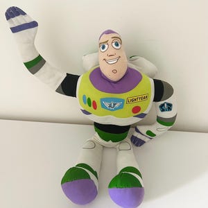 May include: A plush Buzz Lightyear doll from the Toy Story franchise. The doll features a white and green spacesuit, a purple head, and a smiling face. The doll's arm is raised in a waving gesture.
