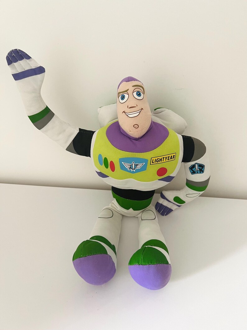 May include: A plush Buzz Lightyear doll from the Toy Story franchise. The doll features a white and green spacesuit, a purple head, and a smiling face. The doll's arm is raised in a waving gesture.