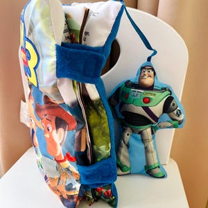 May include: A soft fabric Toy Story 3 book with a blue velcro strap and a plush Buzz Lightyear character attached by a blue ribbon. The book cover features Woody and the text Toy Story 3. The pages are visible inside the soft, padded cover.