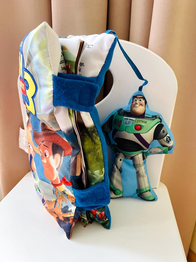 May include: A soft fabric Toy Story 3 book with a blue velcro strap and a plush Buzz Lightyear character attached by a blue ribbon. The book cover features Woody and the text Toy Story 3. The pages are visible inside the soft, padded cover.
