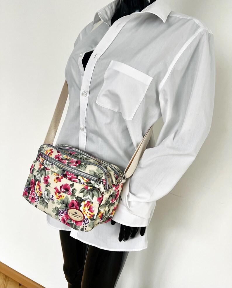 Op de afbeelding: Crossbody tas met bloemenprint op een cr&egrave;mekleurige achtergrond, met rode, roze en gele rozen. De tas heeft een beige band en zilveren ritsen. De tas heeft een klein label met tekst.