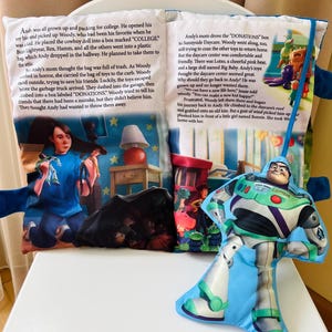 May include: A soft fabric storybook featuring Toy Story scenes and text, accompanied by a plush Buzz Lightyear character attached by a blue ribbon. The pages display illustrations of Andy, Woody, and other toys, with printed narrative text about college and donations.