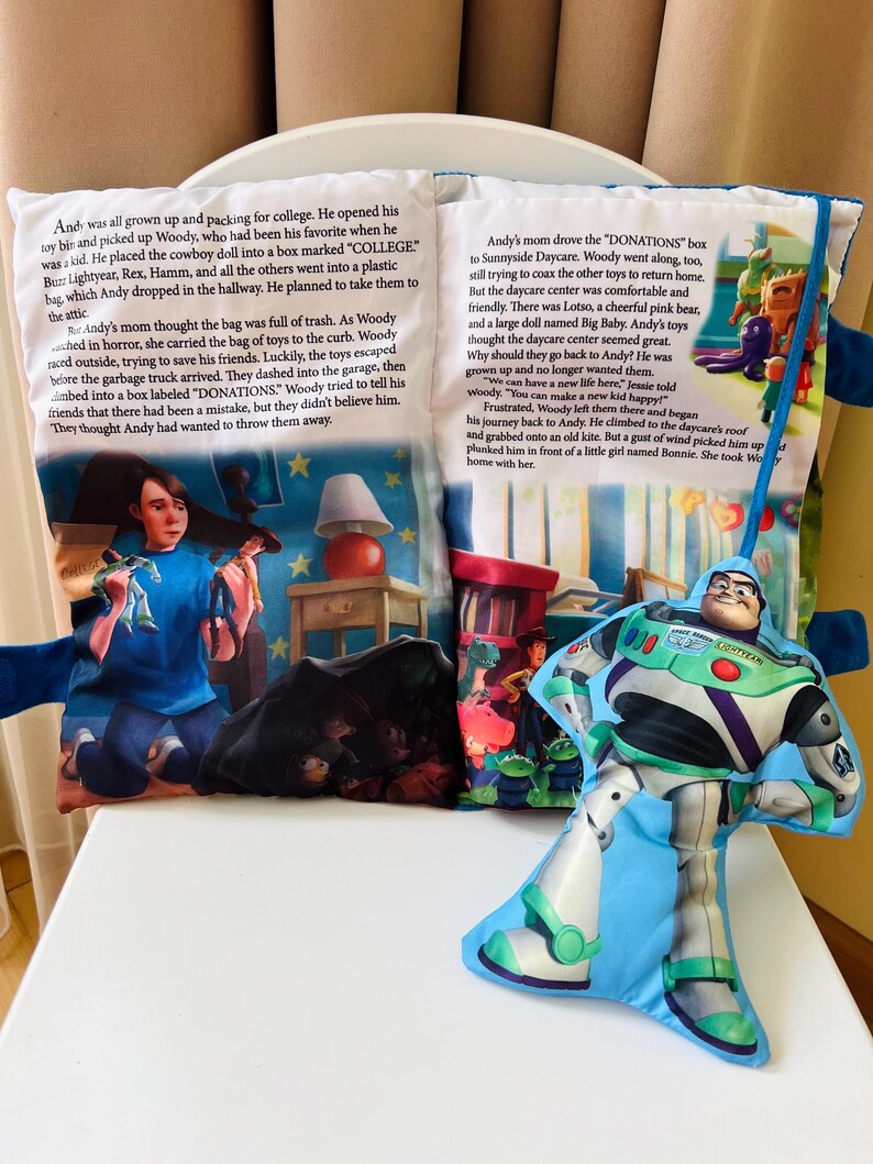 May include: A soft fabric storybook featuring Toy Story scenes and text, accompanied by a plush Buzz Lightyear character attached by a blue ribbon. The pages display illustrations of Andy, Woody, and other toys, with printed narrative text about college and donations.