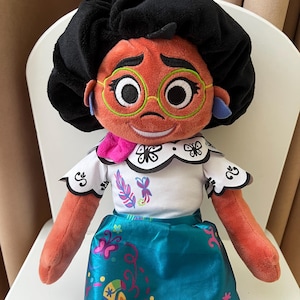 Può includere: Bambola di peluche di un personaggio con pelle arancione, capelli neri e occhiali rotondi verdi. La bambola indossa un top bianco con finiture nere e una gonna turchese con motivi floreali colorati. Una sciarpa rosa è intorno al collo.
