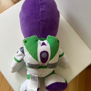 Pode incluir: Um brinquedo de pel&uacute;cia Buzz Lightyear, visto por tr&aacute;s, com uma cabe&ccedil;a roxa e detalhes brancos, verdes e roxos. O brinquedo tem um design de mochila com asas verdes e brancas. Os p&eacute;s s&atilde;o roxos.