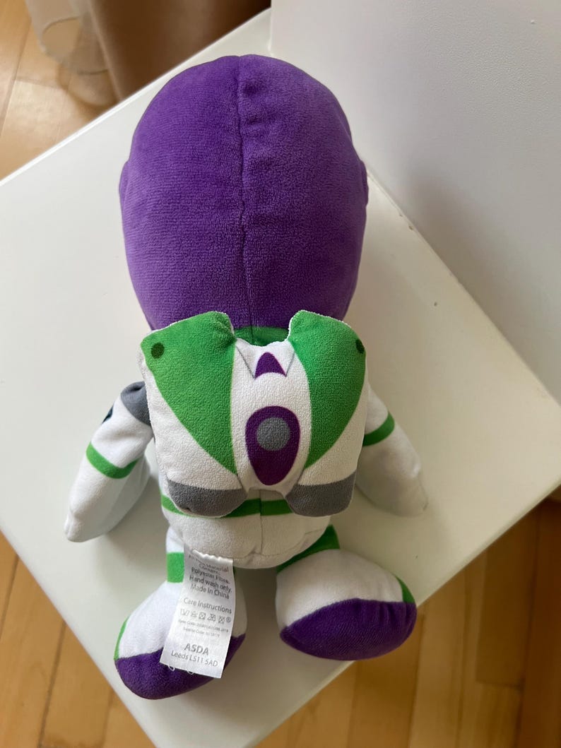 Pode incluir: Um brinquedo de pel&uacute;cia Buzz Lightyear, visto por tr&aacute;s, com uma cabe&ccedil;a roxa e detalhes brancos, verdes e roxos. O brinquedo tem um design de mochila com asas verdes e brancas. Os p&eacute;s s&atilde;o roxos.