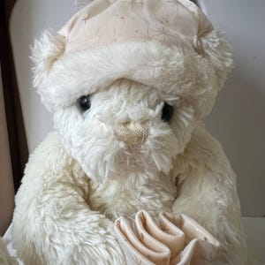 May include: A cream-colored teddy bear wearing a matching hat with a star pattern. The plush bear has black eyes and a detailed nose. It holds a light pink gift bag with a silver ribbon. The bear is a soft, cuddly toy.