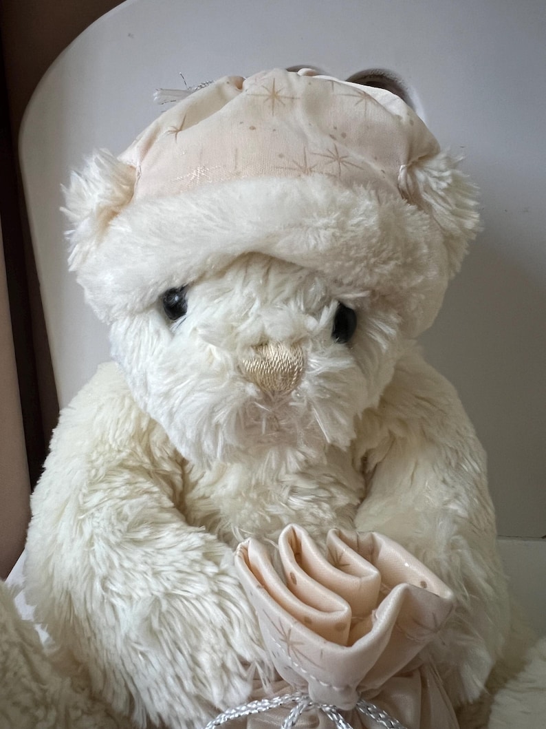May include: A cream-colored teddy bear wearing a matching hat with a star pattern. The plush bear has black eyes and a detailed nose. It holds a light pink gift bag with a silver ribbon. The bear is a soft, cuddly toy.