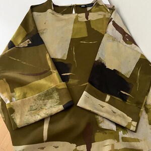 May include: An olive green blouse with a unique abstract pattern in shades of beige, black, and brown. The long sleeves have folded cuffs. The top has a round neckline and a keyhole back.