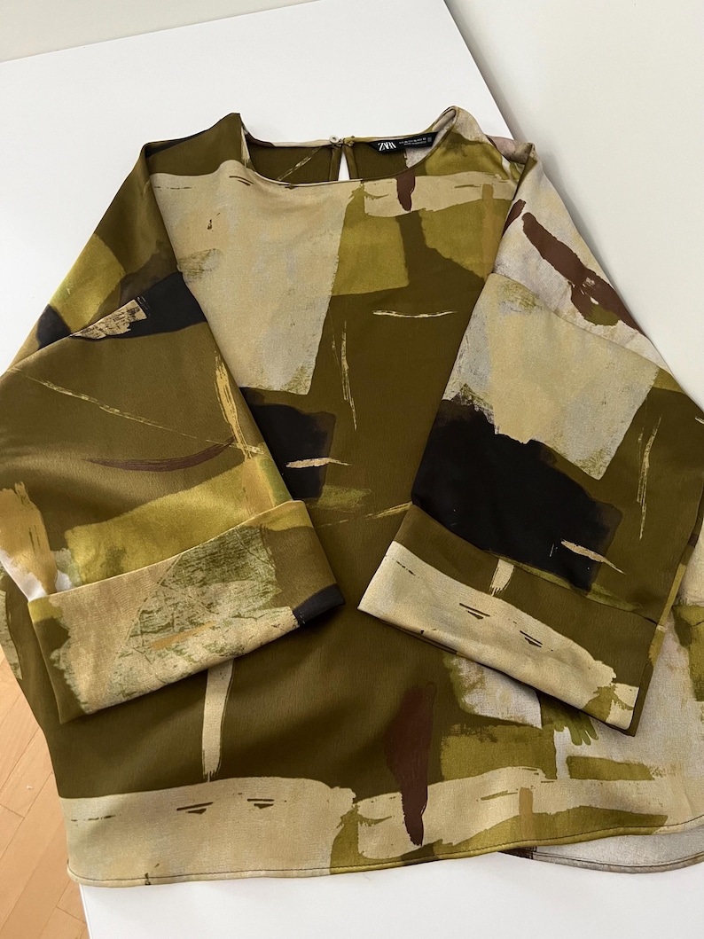 May include: An olive green blouse with a unique abstract pattern in shades of beige, black, and brown. The long sleeves have folded cuffs. The top has a round neckline and a keyhole back.