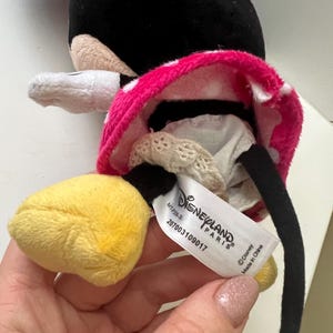May include: A plush Minnie Mouse doll with a black head, white gloves, and yellow feet. The doll wears a pink skirt with white polka dots. A tag reads "Disneyland Paris" and "Made in China."