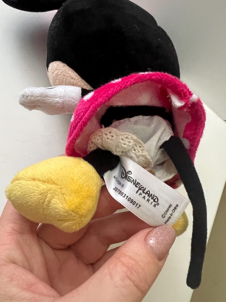 May include: A plush Minnie Mouse doll with a black head, white gloves, and yellow feet. The doll wears a pink skirt with white polka dots. A tag reads "Disneyland Paris" and "Made in China."
