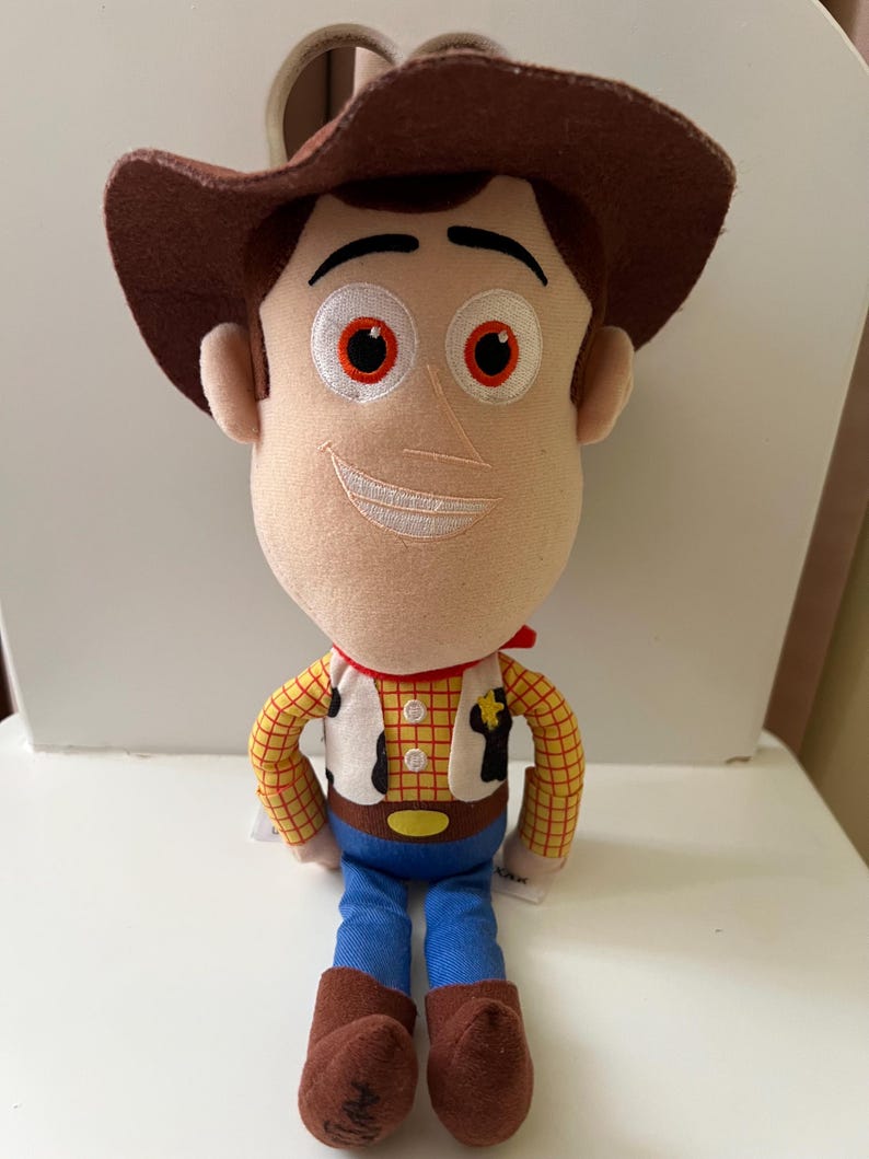 Puede incluir: Un peluche de un personaje de la pel&iacute;cula animada Toy Story. La mu&ntilde;eca tiene un sombrero de vaquero marr&oacute;n, una cara beige con rasgos bordados y un pa&ntilde;uelo rojo. Viste una camisa a cuadros amarilla, un chaleco blanco, vaqueros azules y botas marrones.