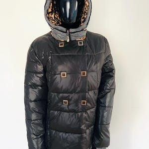 Puede incluir: Abrigo acolchado negro con diseño cruzado y botones cuadrados dorados. El abrigo presenta una capucha gris con forro de estampado de leopardo y un cuello gris. Las mangas tienen puños grises.