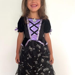 May include: A child's Halloween costume featuring a black velvet bodice with a lavender panel and silver trim. The dress has a black skirt with a spider and star pattern. The costume includes short sleeves with lavender bows.