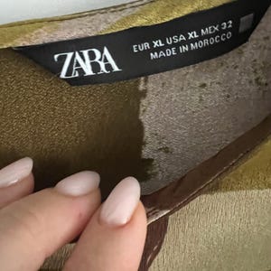 May include: Close-up of a garment label with the brand name "ZABA" in white letters. The label also includes size information: EUR XL, USA XL, MEX 32, and the text "MADE IN MOROCCO". The fabric has a green and brown pattern.