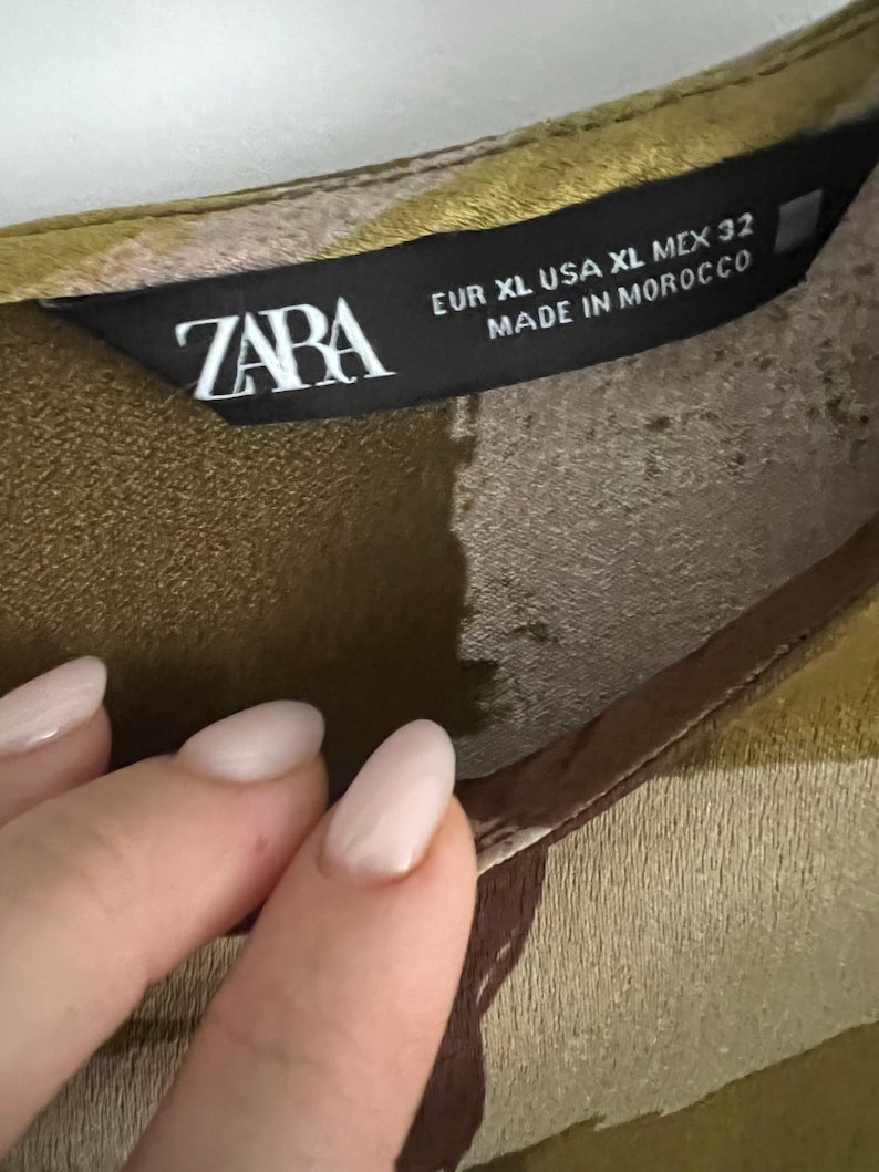 May include: Close-up of a garment label with the brand name "ZABA" in white letters. The label also includes size information: EUR XL, USA XL, MEX 32, and the text "MADE IN MOROCCO". The fabric has a green and brown pattern.