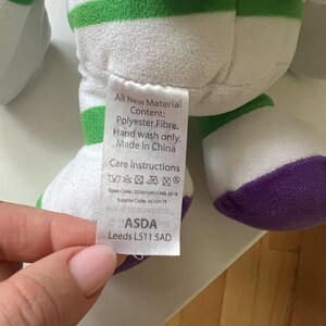 Pode incluir: Um brinquedo de pel&uacute;cia em branco, verde, roxo e cinza. O brinquedo tem listras verdes e detalhes roxos. Uma etiqueta com instru&ccedil;&otilde;es de cuidado e o texto "All New Material Content: Polyester Fibre. Hand wash only. Made in China" &eacute; vis&iacute;vel.