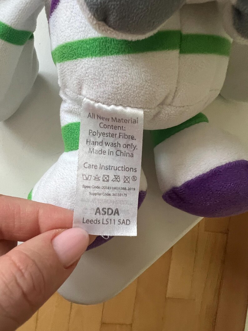 Pode incluir: Um brinquedo de pel&uacute;cia em branco, verde, roxo e cinza. O brinquedo tem listras verdes e detalhes roxos. Uma etiqueta com instru&ccedil;&otilde;es de cuidado e o texto "All New Material Content: Polyester Fibre. Hand wash only. Made in China" &eacute; vis&iacute;vel.