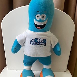 Może przedstawiać: Niebieska pluszowa zabawka w kształcie uśmiechniętej postaci, nosząca białą koszulkę i niebieskie szorty. Na koszulce widnieje napis "Maths of the Day" i "Active Maths". Stopy są niebieskie, białe i pomarańczowe.