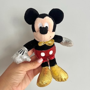 Puede incluir: Un peluche de Mickey Mouse con cabeza y orejas negras, cara color canela y una pajarita dorada brillante. El personaje lleva pantalones cortos rojos brillantes con dos botones blancos, guantes plateados brillantes y zapatos dorados brillantes.
