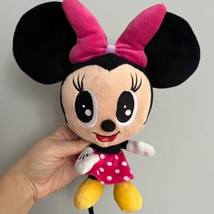 May include: A plush Minnie Mouse doll with black ears, a pink bow, and a pink and white polka dot dress. The doll has a smiling face with large black eyes and yellow shoes. The doll is a classic Disney character.