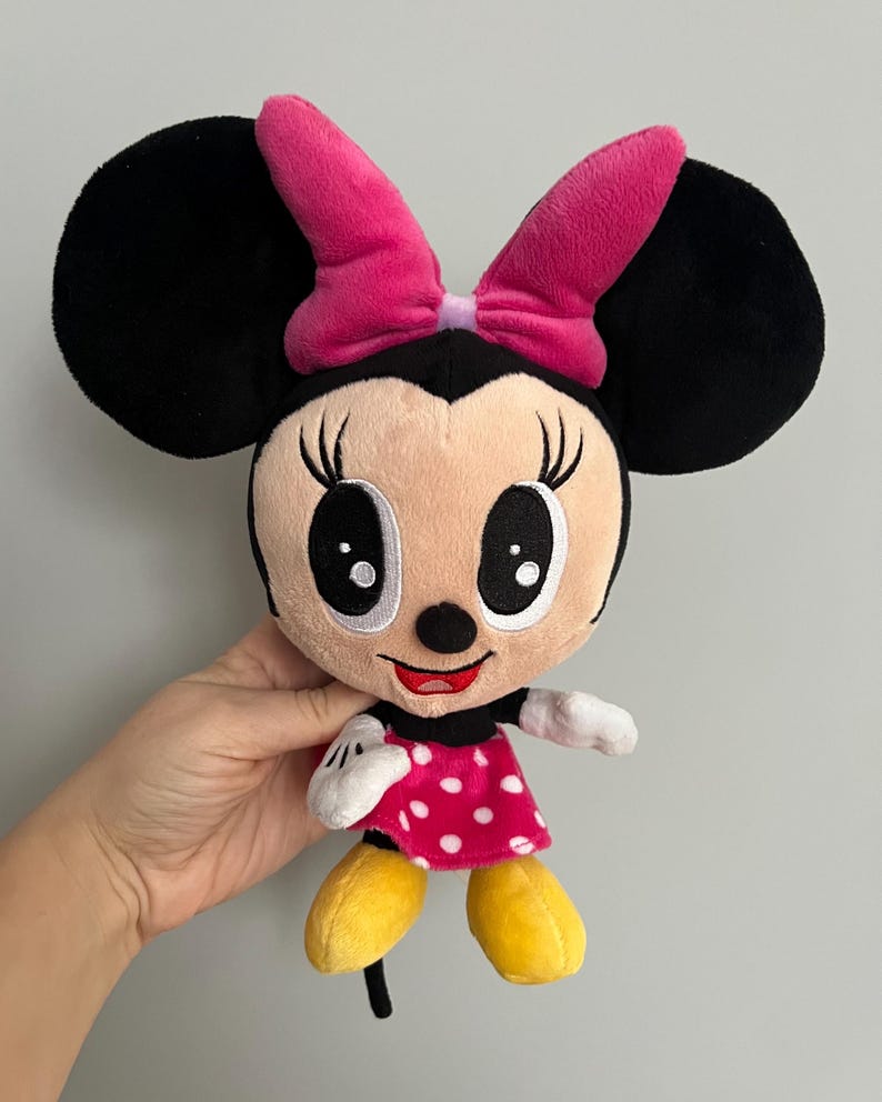May include: A plush Minnie Mouse doll with black ears, a pink bow, and a pink and white polka dot dress. The doll has a smiling face with large black eyes and yellow shoes. The doll is a classic Disney character.