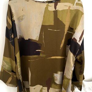 May include: An abstract print blouse in shades of olive green, beige, brown, and black. The long-sleeved top features a round neckline and wide cuffs. The fabric has a silky sheen, and the design is reminiscent of an artistic painting.