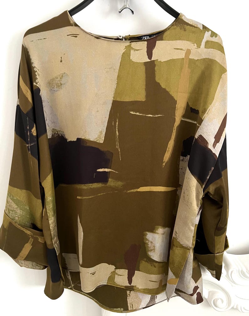 May include: An abstract print blouse in shades of olive green, beige, brown, and black. The long-sleeved top features a round neckline and wide cuffs. The fabric has a silky sheen, and the design is reminiscent of an artistic painting.