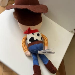 Puede incluir: Mu&ntilde;eco de peluche de Woody de la franquicia Disney Pixar Toy Story. El mu&ntilde;eco presenta un sombrero de vaquero marr&oacute;n, una camisa a cuadros amarilla, vaqueros azules y botas marrones. Una bandana roja est&aacute; atada alrededor del cuello. La etiqueta de Disney Pixar es visible.