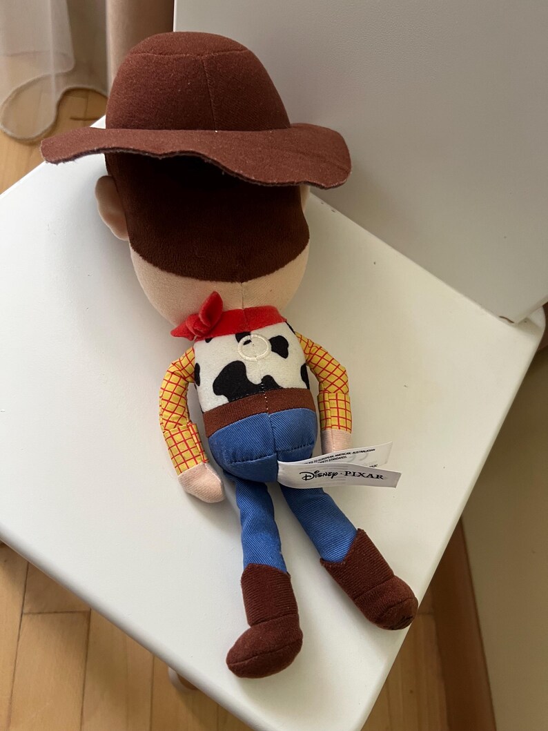 Puede incluir: Mu&ntilde;eco de peluche de Woody de la franquicia Disney Pixar Toy Story. El mu&ntilde;eco presenta un sombrero de vaquero marr&oacute;n, una camisa a cuadros amarilla, vaqueros azules y botas marrones. Una bandana roja est&aacute; atada alrededor del cuello. La etiqueta de Disney Pixar es visible.