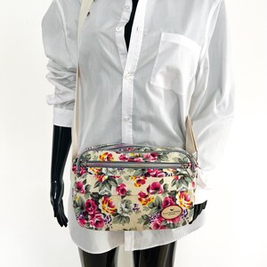 Op de afbeelding: Een bloemen crossbodytas met een cr&egrave;mekleurige achtergrond en een patroon van roze, rode en gele rozen. De tas heeft een grijze rits en een cr&egrave;mekleurige band. Een klein, bruin leren label is bevestigd.
