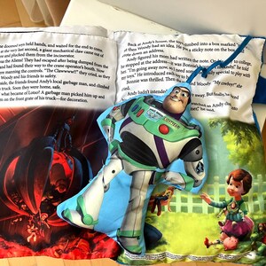 May include: A soft fabric book featuring a printed story about toys and a plush Buzz Lightyear character attached to the page. The pages display colorful illustrations of a mechanical claw and a child playing, alongside printed text in English.