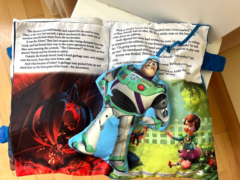 May include: A soft fabric book featuring a printed story about toys and a plush Buzz Lightyear character attached to the page. The pages display colorful illustrations of a mechanical claw and a child playing, alongside printed text in English.