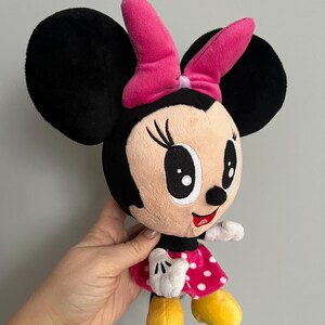 May include: A plush Minnie Mouse doll with large black ears, a pink bow, and a red polka-dot dress. The doll has white gloves and yellow shoes. A classic Disney character.
