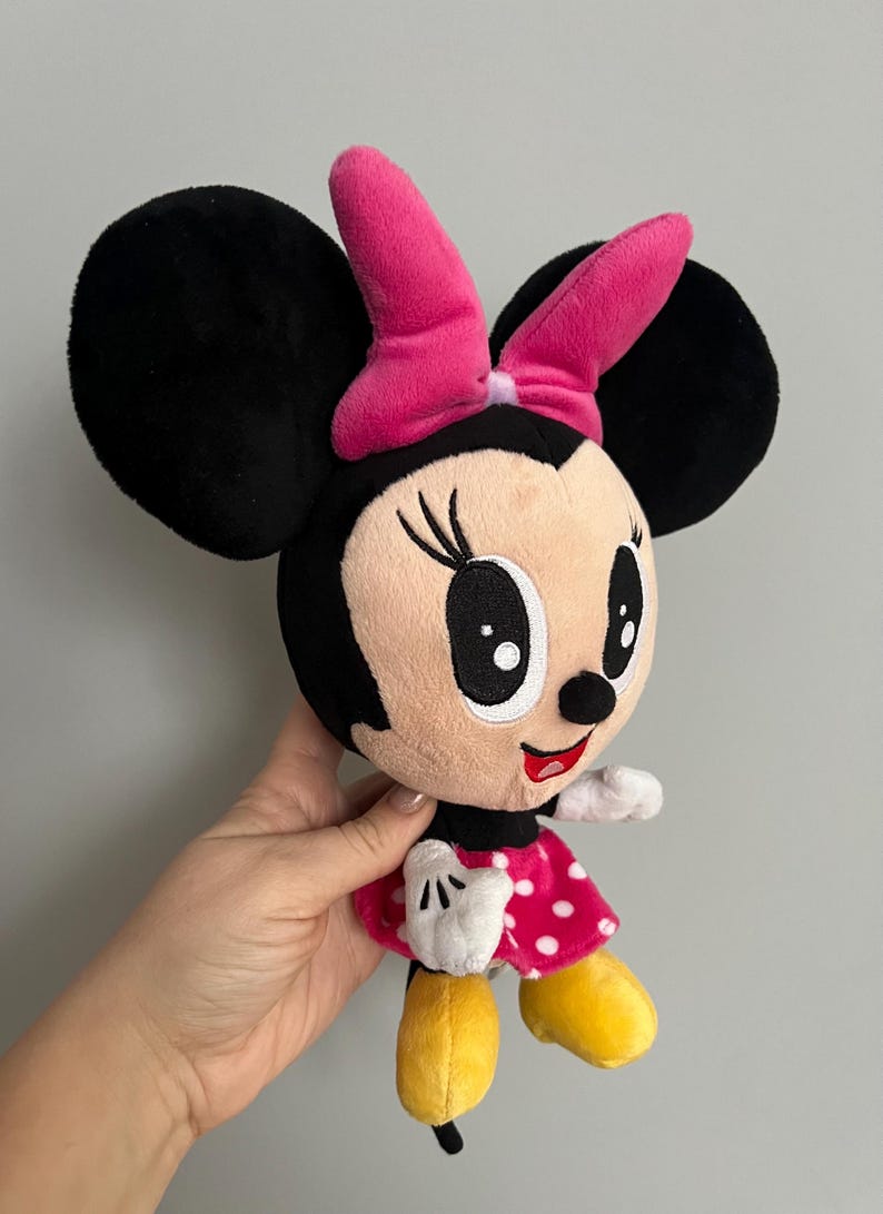 May include: A plush Minnie Mouse doll with large black ears, a pink bow, and a red polka-dot dress. The doll has white gloves and yellow shoes. A classic Disney character.