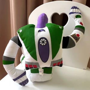 May include: A plush toy resembling the character Buzz Lightyear from the animated film Toy Story. The toy features a white body with green and purple accents, including a helmet, wings, and a circular emblem on the chest. The arms are outstretched, and the toy is sitting on a white chair.