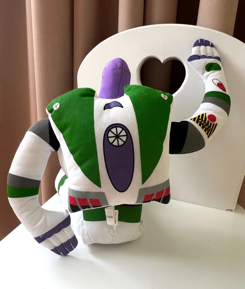 May include: A plush toy resembling the character Buzz Lightyear from the animated film Toy Story. The toy features a white body with green and purple accents, including a helmet, wings, and a circular emblem on the chest. The arms are outstretched, and the toy is sitting on a white chair.