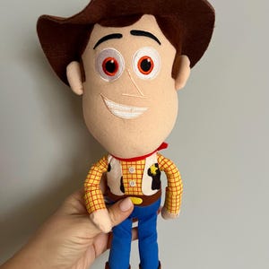 Puede incluir: Un peluche de un personaje de dibujos animados de vaquero. La mu&ntilde;eca tiene un sombrero marr&oacute;n, una cara beige con ojos rojos y una sonrisa cosida. Viste una camisa a cuadros amarillos, un chaleco, pantalones azules y botas marrones.