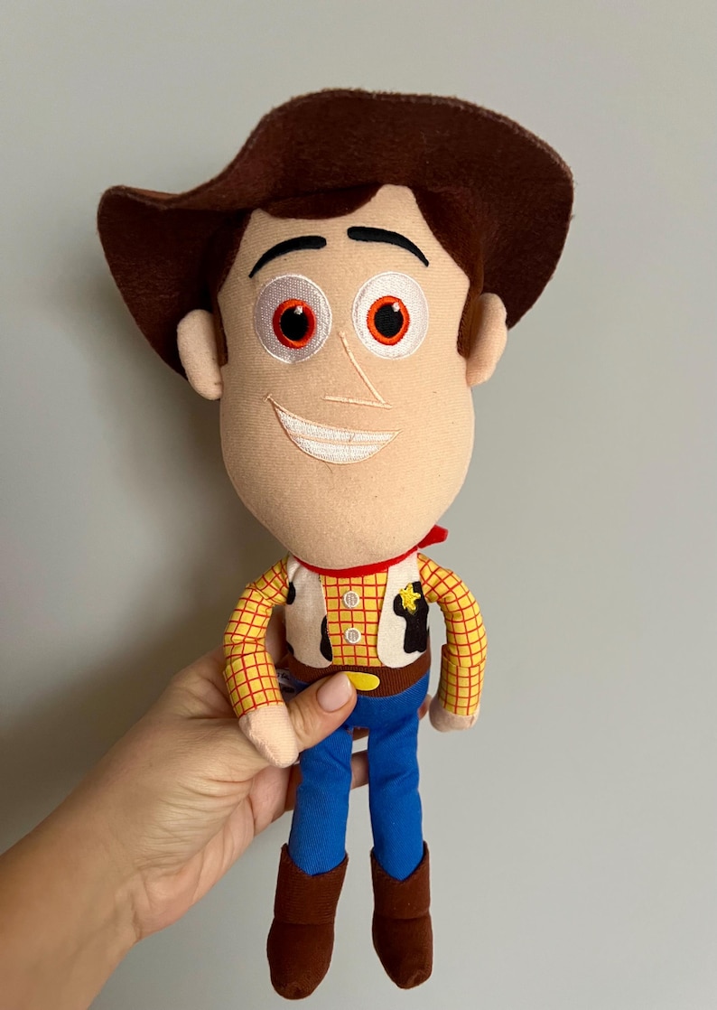 Puede incluir: Un peluche de un personaje de dibujos animados de vaquero. La mu&ntilde;eca tiene un sombrero marr&oacute;n, una cara beige con ojos rojos y una sonrisa cosida. Viste una camisa a cuadros amarillos, un chaleco, pantalones azules y botas marrones.
