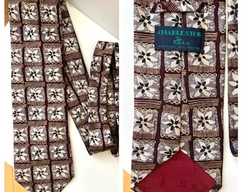 Op de afbeelding: Een stropdas met een patroon met een herhalend bloemmotief in tinten bordeauxrood, grijs en crème. De stropdas heeft een vierkant patroon met een bloemmotief. Het label luidt "Charleston Tie Rack".