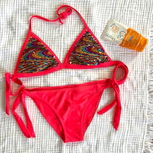 Op de afbeelding: Een felroze bikiniset met een driehoekige top en strikzijden. De top heeft kleurrijke pailletten details. Er is ook een tube zonnebrandcrème met SPF 30 bescherming aanwezig.