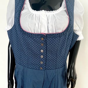 Könnte beinhalten: Ein traditionelles blaues Dirndlkleid mit einer weißen Bluse. Das Kleid hat ein gemustertes Oberteil mit dekorativen Knöpfen und einen gepunkteten Rock. Der Ausschnitt ist mit rosa und weißer Spitze verziert.