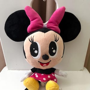May include: A plush Minnie Mouse doll with black ears, a pink bow, and a pink and white polka dot dress. The doll has a smiling face with large eyes and yellow shoes. The doll is sitting on a white chair.