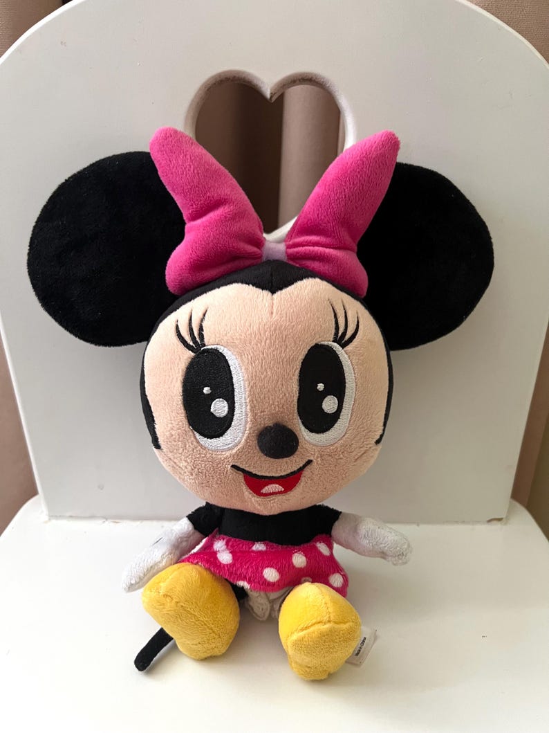 May include: A plush Minnie Mouse doll with black ears, a pink bow, and a pink and white polka dot dress. The doll has a smiling face with large eyes and yellow shoes. The doll is sitting on a white chair.