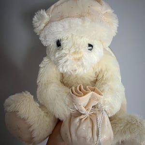 May include: A cream-colored teddy bear wearing a matching hat with gold starburst patterns. The plush bear holds a small gift bag with a silver drawstring. The bear's paws and hat feature the same starburst design.